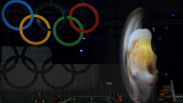 Eine Turnerin führt vor dem Hintergrund der olympischen Ringe eine Übung auf dem Trampolin aus.