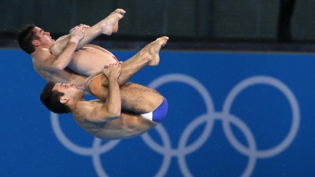 Zwei Wasserspringer führen einen synchronen Sprung vor dem Hintergrund der olympischen Ringe aus.