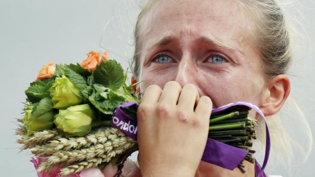 Eine weinende Frau hält einen Blumenstrauß mit einer lila Schleife vor ihr Gesicht.