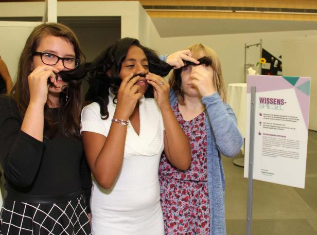 Drei junge Frauen halten ihre Haare wie Schnurrbärte unter die Nase.