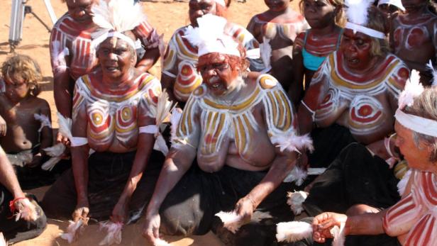 Eine Gruppe indigener Australier mit traditioneller Körperbemalung und Federschmuck sitzt auf dem Boden.