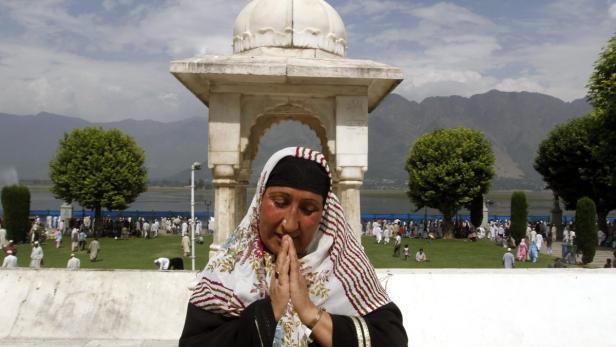 Eine muslimische Frau betet vor einem Schrein mit Blick auf eine Menschenmenge und Berge.