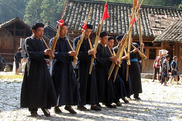 Eine Gruppe von Männern in traditioneller Kleidung spielt Bambusinstrumente in einem Dorf.