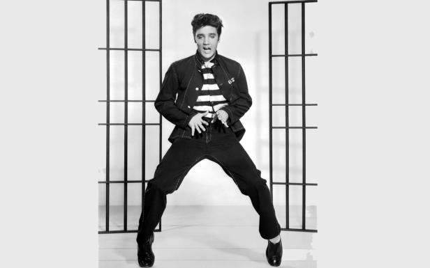 Elvis Presley in einem gestreiften Hemd und einer dunklen Jacke vor einem Gitter.