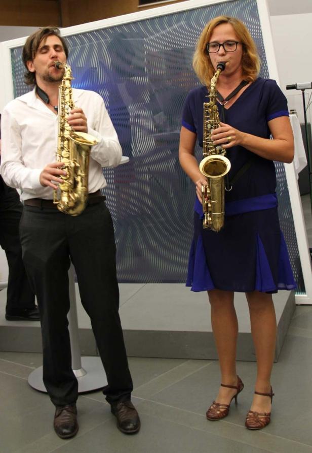 Ein Mann und eine Frau spielen Saxophon vor einem abstrakten Hintergrund.