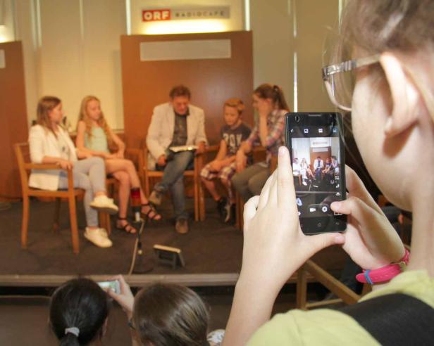 Ein Mädchen fotografiert mit ihrem Smartphone eine Gruppe von Personen auf einer Bühne.