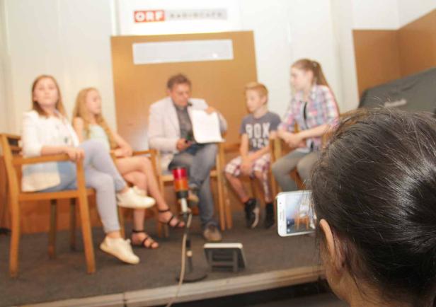Eine Gruppe von Kindern und ein Mann sitzen auf Stühlen vor einem ORF-Logo.