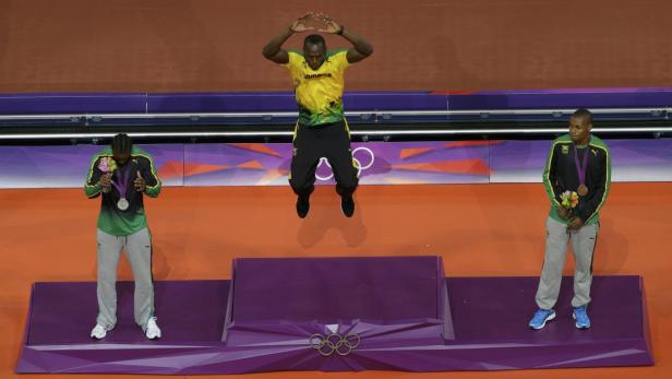Usain Bolt springt auf dem Siegerpodest, flankiert von zwei weiteren jamaikanischen Athleten mit Medaillen.