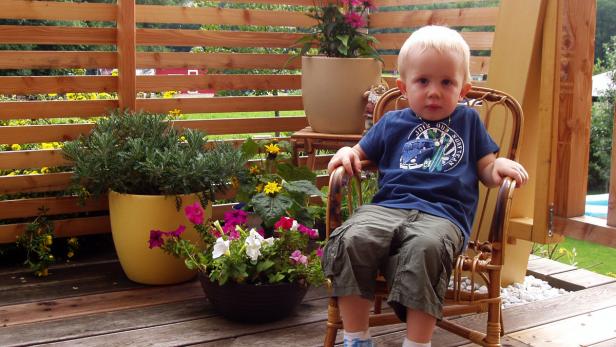 Ein kleiner Junge sitzt auf einem Rattanstuhl auf einer Terrasse mit Blumen.