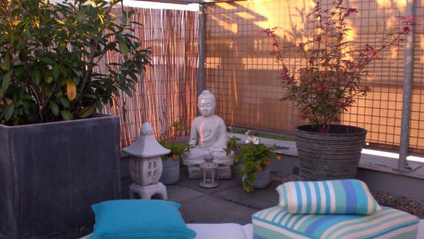 Eine Buddha-Statue sitzt inmitten von Pflanzen und Kissen auf einer Terrasse.
