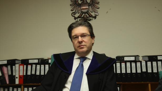 Richter Georg Olschak. Ein Mann in Robe und Krawatte sitzt vor einem Regal mit Aktenordnern und dem österreichischen Bundesadler.