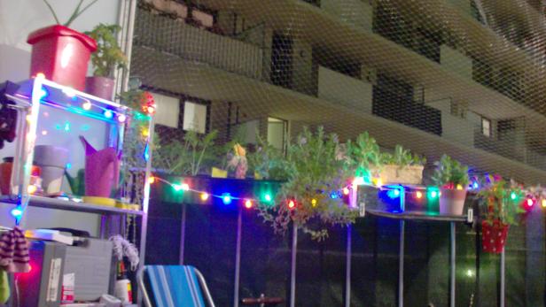 Ein mit bunten Lichtern geschmückter Balkon mit Pflanzen und Gartenutensilien.