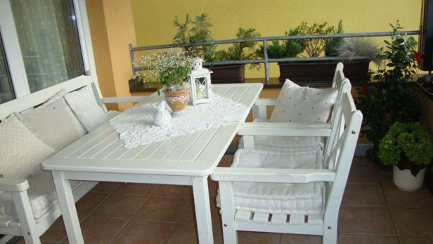 Ein weißer Tisch und Stühle stehen auf einem sonnigen Balkon mit Pflanzen.