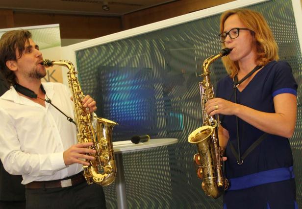 Zwei Personen spielen Saxophon vor einem gemusterten Hintergrund.