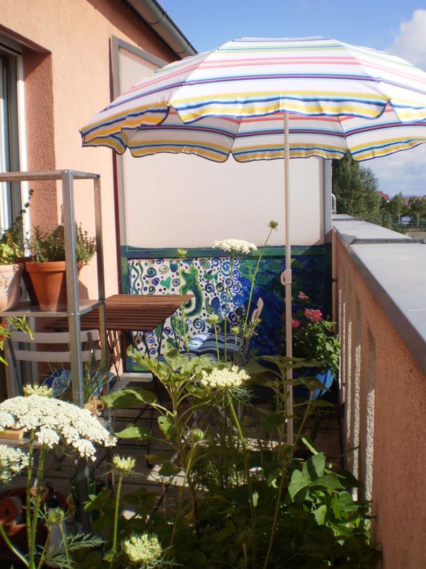 Ein sonniger Balkon mit einem gestreiften Sonnenschirm und blühenden Pflanzen.