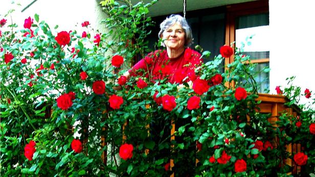Eine ältere Frau lächelt hinter einem Rosenbusch auf einem Balkon.