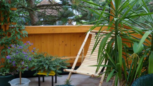 Ein Bambus-Liegestuhl steht auf einer Terrasse mit Pflanzen und einem Holzzaun im Hintergrund.