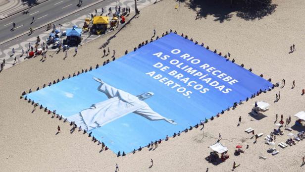 Ein riesiges Banner am Strand von Rio de Janeiro zeigt die Christusstatue und den Text „O Rio recebe as Olimpíadas de braços abertos.“.