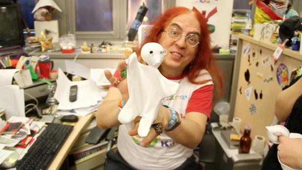 Ein Mann mit rotem Haar hält eine weiße Stoffpuppe mit Brille in die Kamera.