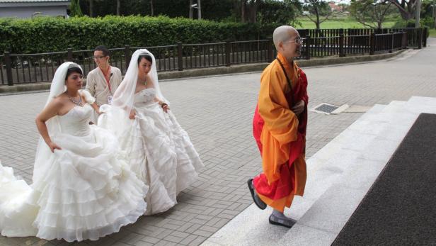 Zwei Frauen in Brautkleidern gehen neben einer buddhistischen Nonne.