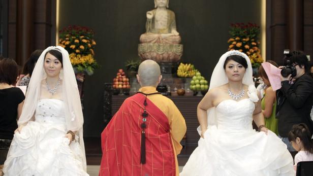 Zwei Bräute in weißen Kleidern stehen neben einem buddhistischen Mönch vor einer Buddha-Statue.