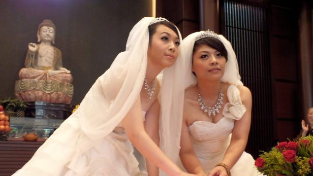 Zwei Frauen in Brautkleidern mit Schleiern und Diademen vor einer Buddha-Statue.