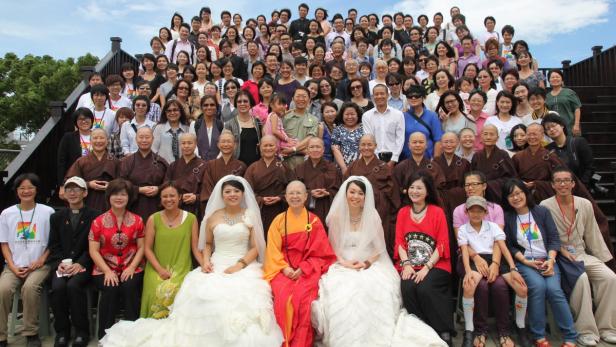 Eine große Gruppe von Menschen, darunter mehrere Paare in Hochzeitskleidern, posiert für ein Foto.