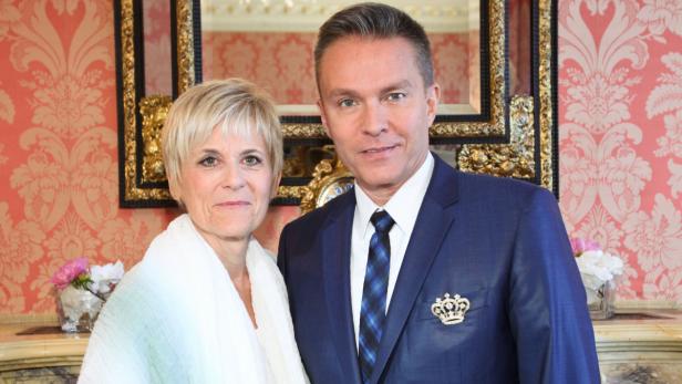 Eine Frau mit einem weißen Tuch und ein Mann im Anzug mit einer Brosche in Kronenform posieren für ein Foto.