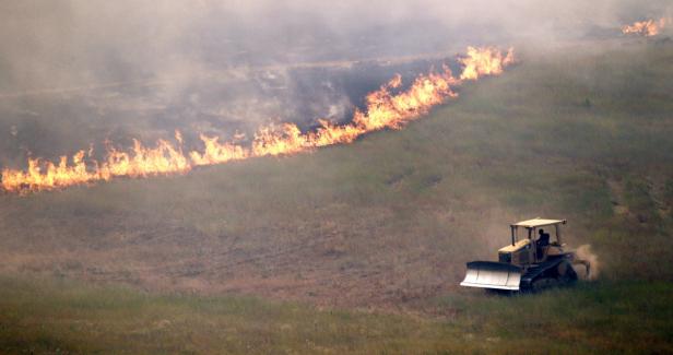 Ein Bulldozer arbeitet an einem Hang, während im Hintergrund ein Feuer brennt.