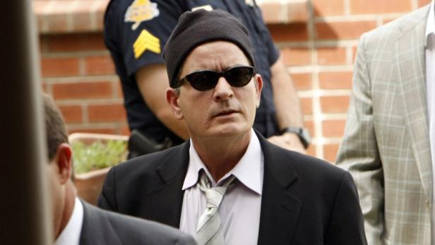 Charlie Sheen mit Sonnenbrille und Mütze, flankiert von einem Polizisten und einem Mann im Anzug.