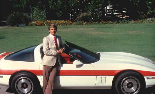 Ein Mann im Anzug posiert neben einem weißen Sportwagen mit rotem Streifen.