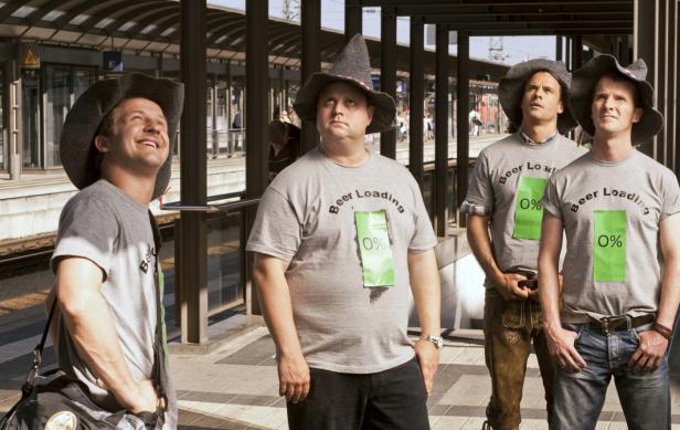 Vier Männer mit Hüten und T-Shirts mit der Aufschrift „Beer Loading 0%“ stehen an einem Bahnhof.