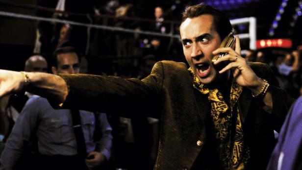 Nicolas Cage gestikuliert aufgeregt mit einem Handy am Ohr.