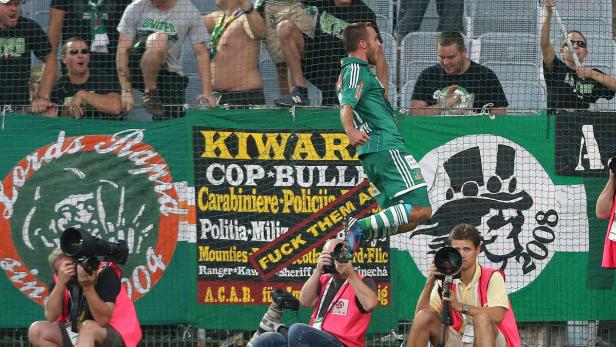 Ein Fußballspieler springt vor einer Zuschauertribüne mit Bannern und Fotografen.