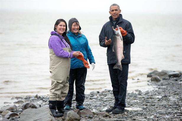 Barack Obama posiert mit zwei Frauen und einem großen Fisch am Ufer.