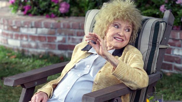 Eine ältere Frau mit blondem Haar sitzt in einem Gartenstuhl und lächelt.