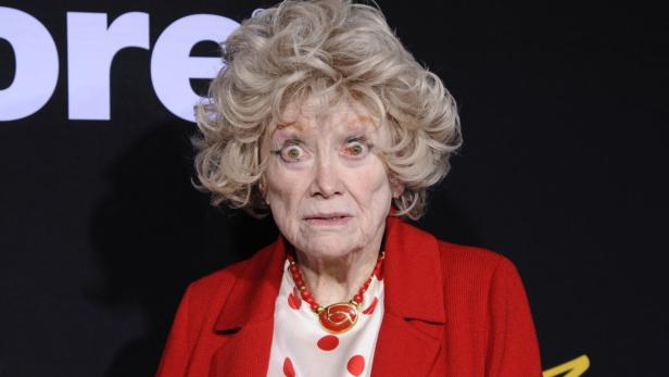 Eine ältere Frau mit blondem Haar, rotem Blazer und auffälligem Make-up blickt in die Kamera.