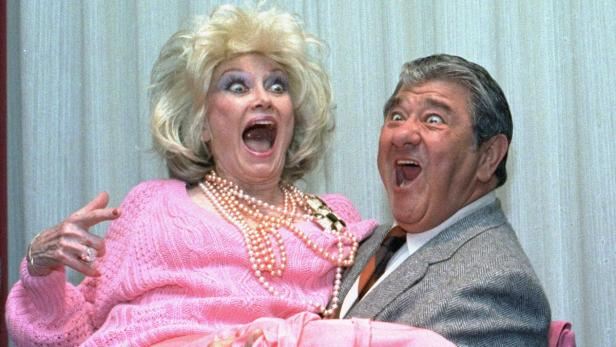 Ein Mann hält eine Frau im Arm, beide mit aufgerissenen Mündern und überraschten Gesichtsausdrücken.