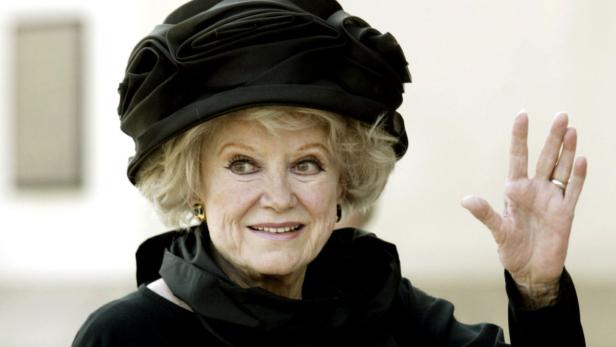 Eine ältere Frau mit Hut winkt in die Kamera.