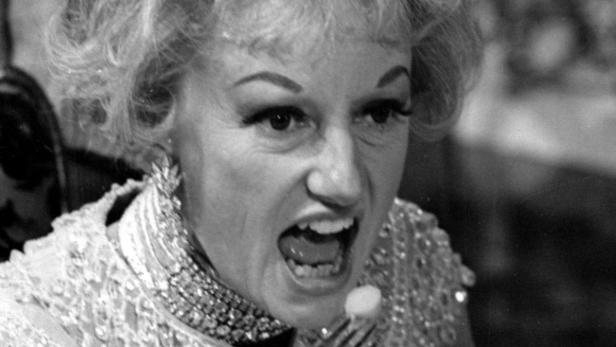 Eine Frau mit blonden Haaren und auffälligem Make-up schreit in ein Mikrofon.