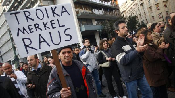 Demonstranten halten ein Schild mit der Aufschrift „Troika Merkel Raus“.