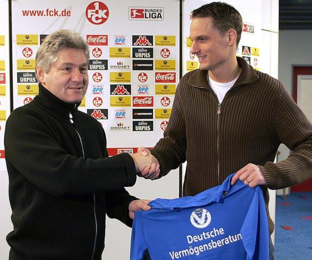 Zwei Männer schütteln sich die Hände vor einer Wand mit Sponsorenlogos.