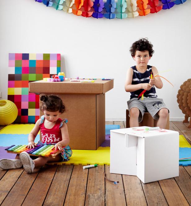 Zwei Kinder spielen in einem Zimmer mit bunten Dekorationen und selbstgebauten Möbeln aus Karton.