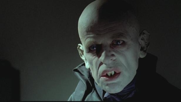 Nahaufnahme eines Mannes mit blasser Haut und spitzen Zähnen, möglicherweise ein Vampir.