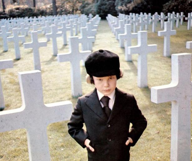 Ein Junge in Anzug und Mütze steht auf einem Soldatenfriedhof.