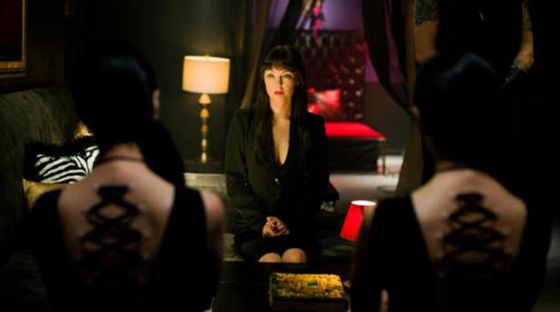 Eine Frau in einem schwarzen Blazer sitzt zwischen zwei Frauen mit Korsett-Tattoos auf dem Rücken.
