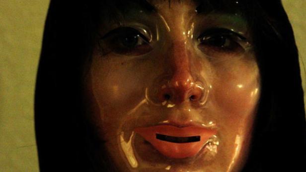 Nahaufnahme einer Frau, die eine transparente Gesichtsmaske mit einem schwarzen Balken über dem Mund trägt.