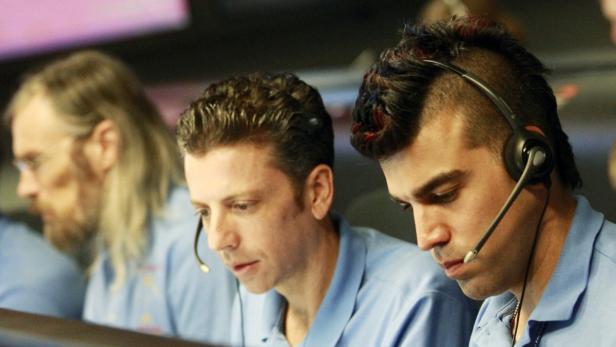 Drei Männer mit Headsets sitzen vor einem Bildschirm.