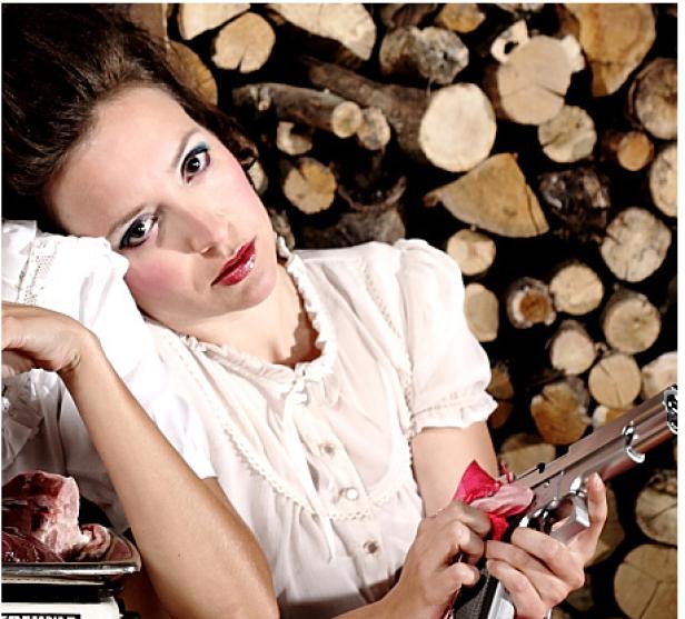 Eine Frau mit blassem Make-up hält eine Pistole in der Hand, neben ihr liegt Fleisch auf einer Waage.