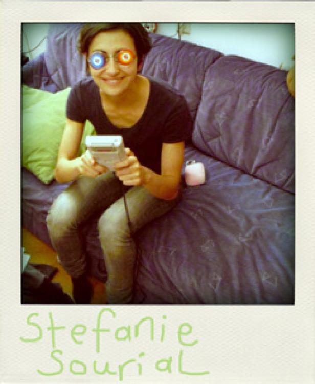 Eine junge Frau mit bunten Augen-Aufklebern spielt auf einem Handheld-Spielgerät, während sie auf einem Sofa sitzt.